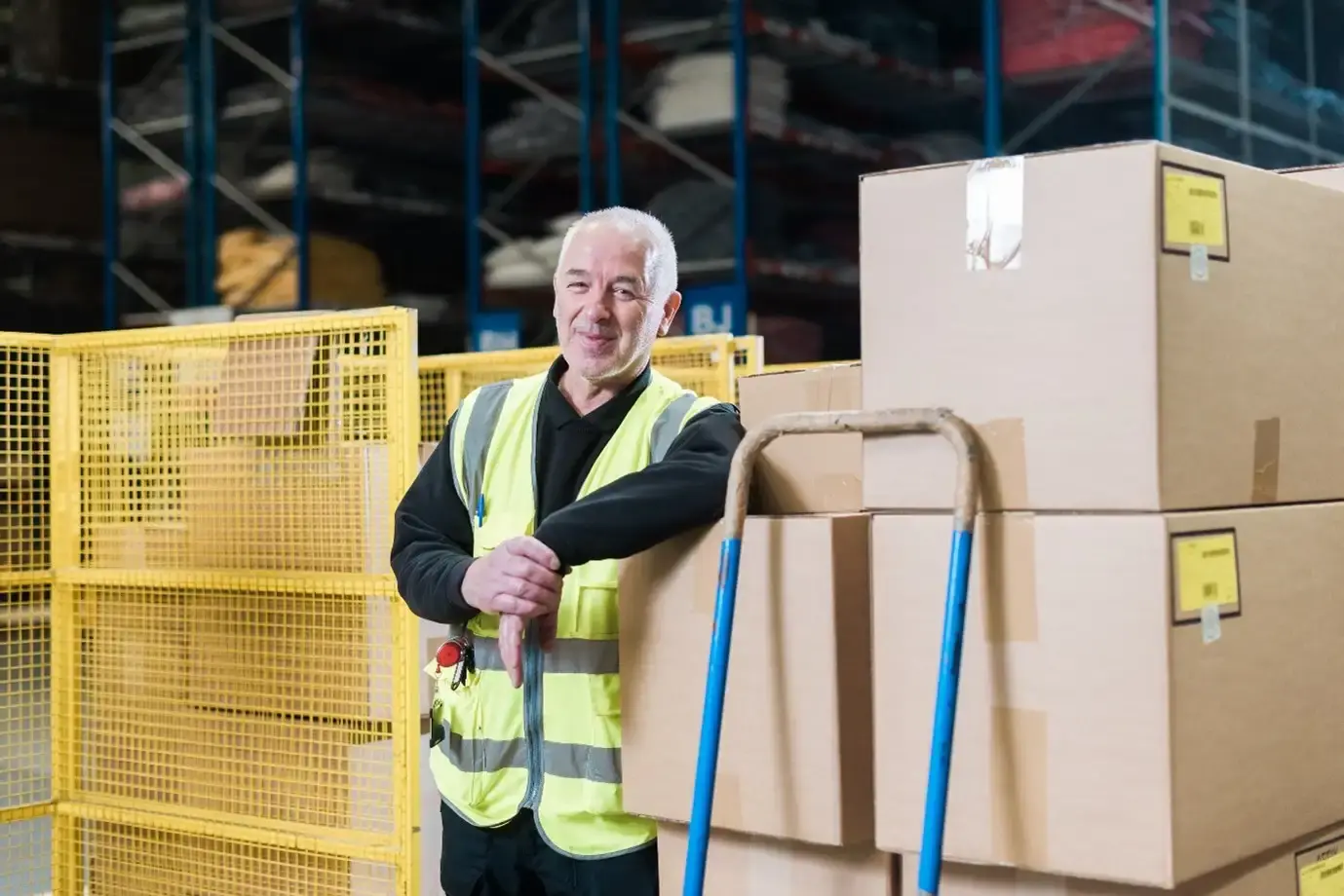 Smiling warehouse worker in a safety vest leans on a stack of large cardboard boxes on a blue-handled cart, with shelves and a cage in the background.